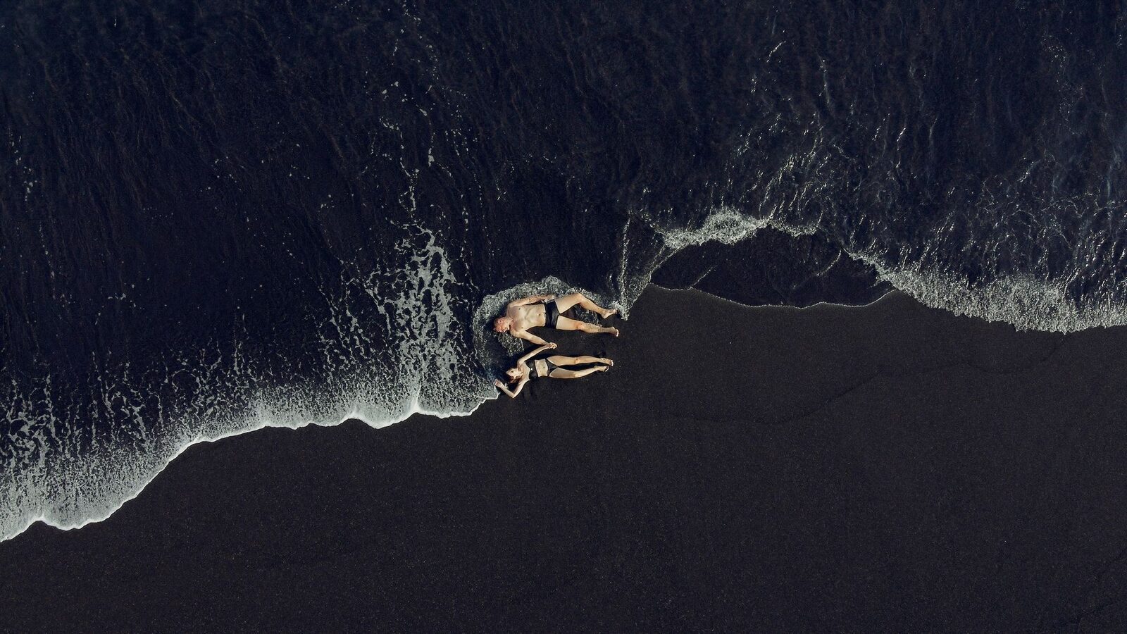 Aerial over the black-sand beach and the line of surf — the volcanic coast from above.