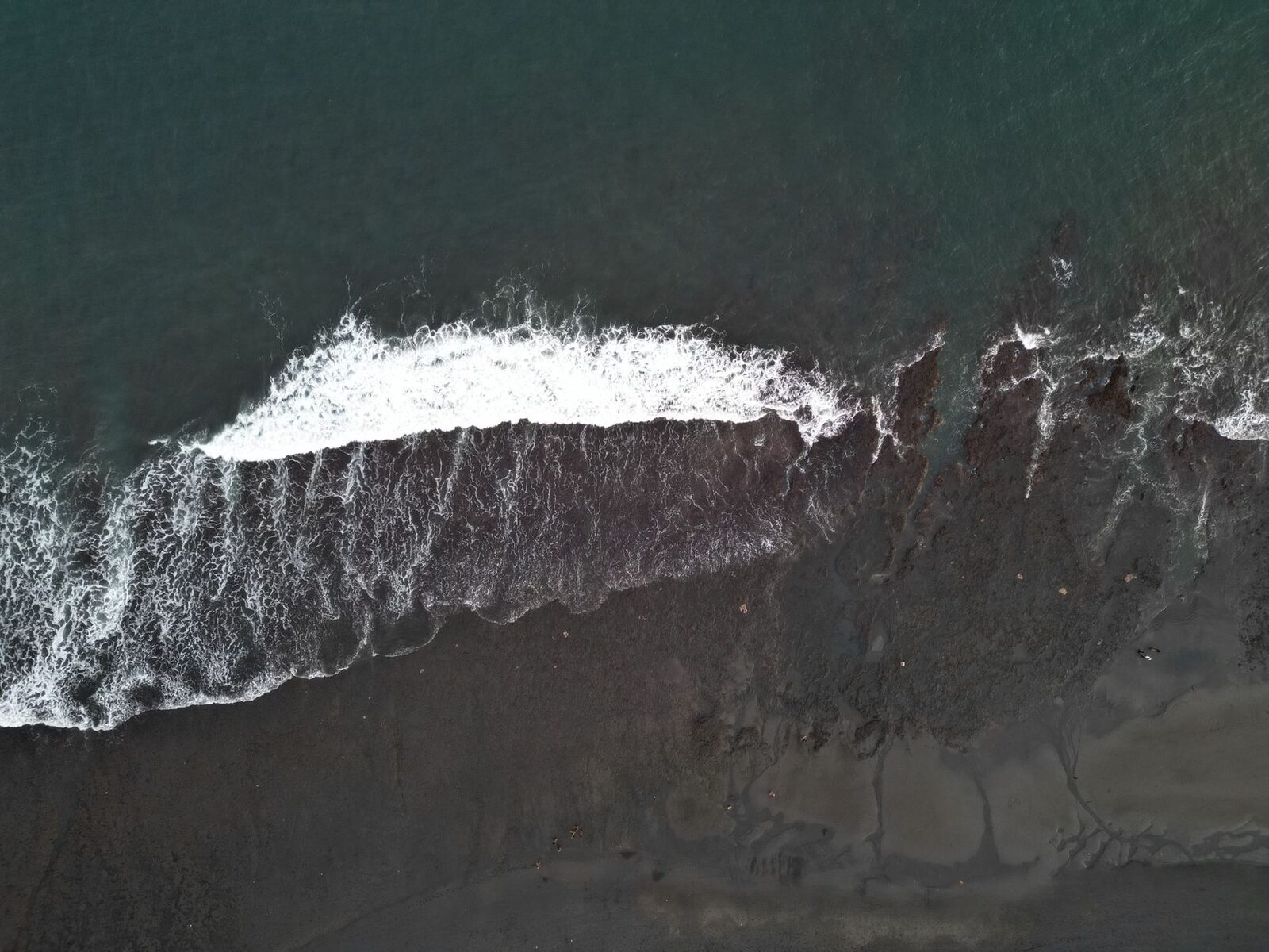 The dark sand and breaking waves seen from the air, austere and elemental.