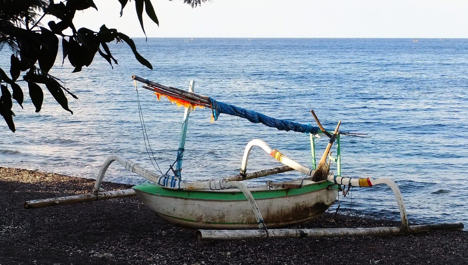 A jukung outrigger hauled up on Amed's black pebble beach, the sea flat behind it — the coast's everyday postcard.