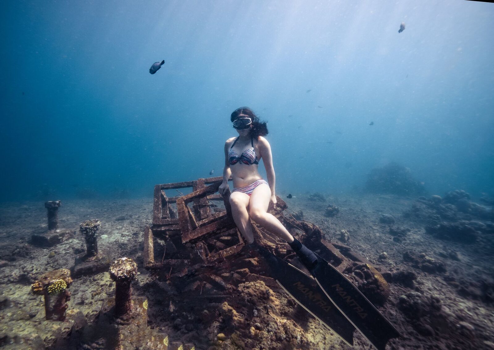 A freediver poised on a submerged structure off Amed, sunlight streaming down through the clear water.