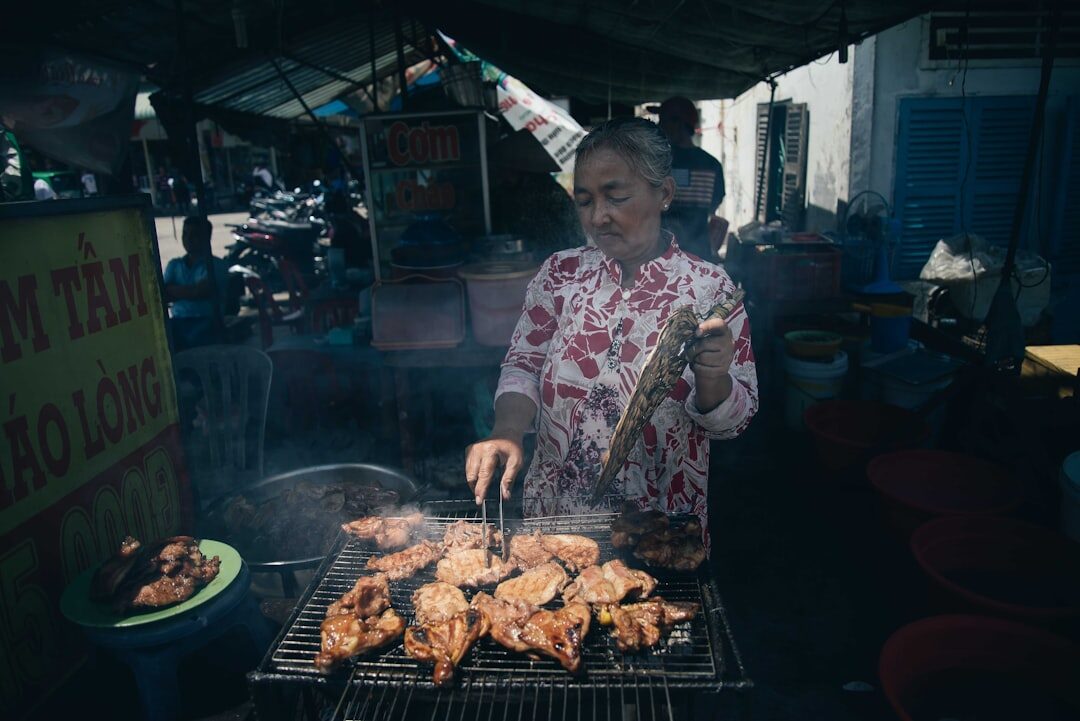 A woman grilling chicken over a smoky fire at a roadside warung.