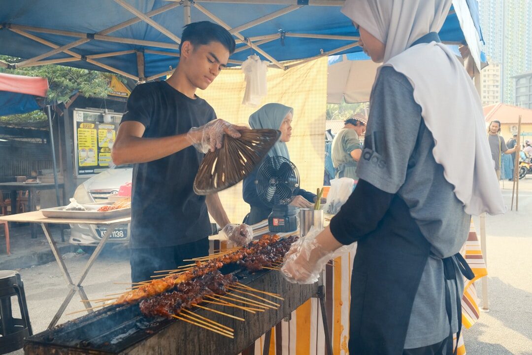 A cook grilling satay to order at a busy street stall, customers waiting — food moving fast and served hot, which is exactly the good sign.