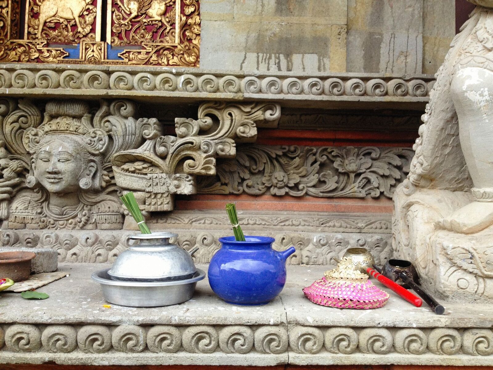 Canang and offerings set along a carved temple ledge beneath a stone guardian.