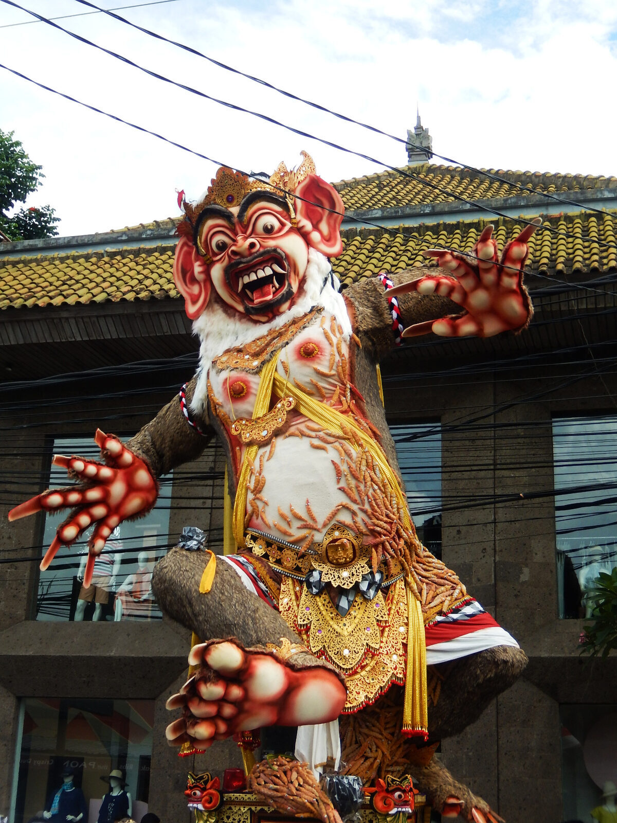 A red, fanged ogoh-ogoh looming over a village street beside a temple gate.