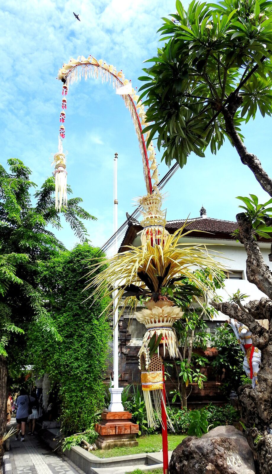 A single penjor curving high over a family gateway, its decorated tip dangling above the entrance.
