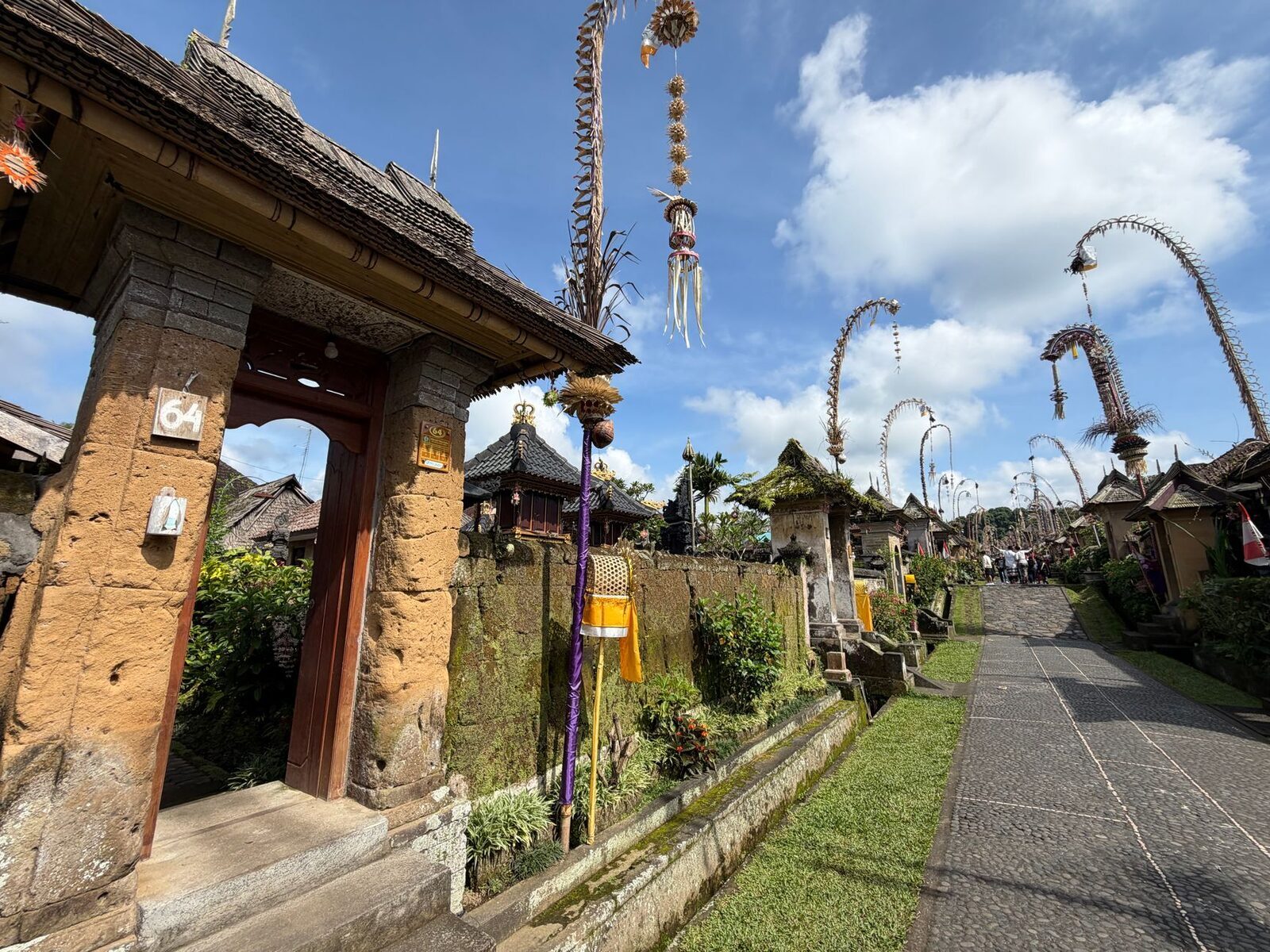 Penjor beside a temple gate and garden, the curved bamboo catching the light.