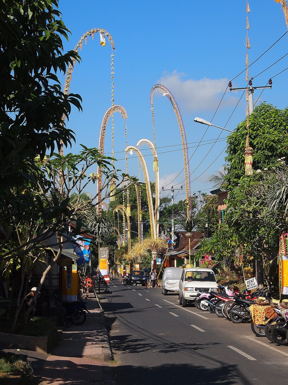 Penjor lining a busier road, the poles leaning together above the traffic.
