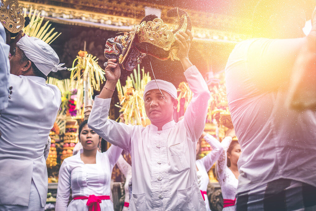 Men in white raising offerings during a temple procession, the sun flaring behind.