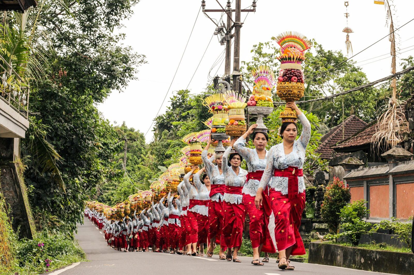 A long line of women in ceremonial dress carrying offerings on their heads down a village road toward the temple.
