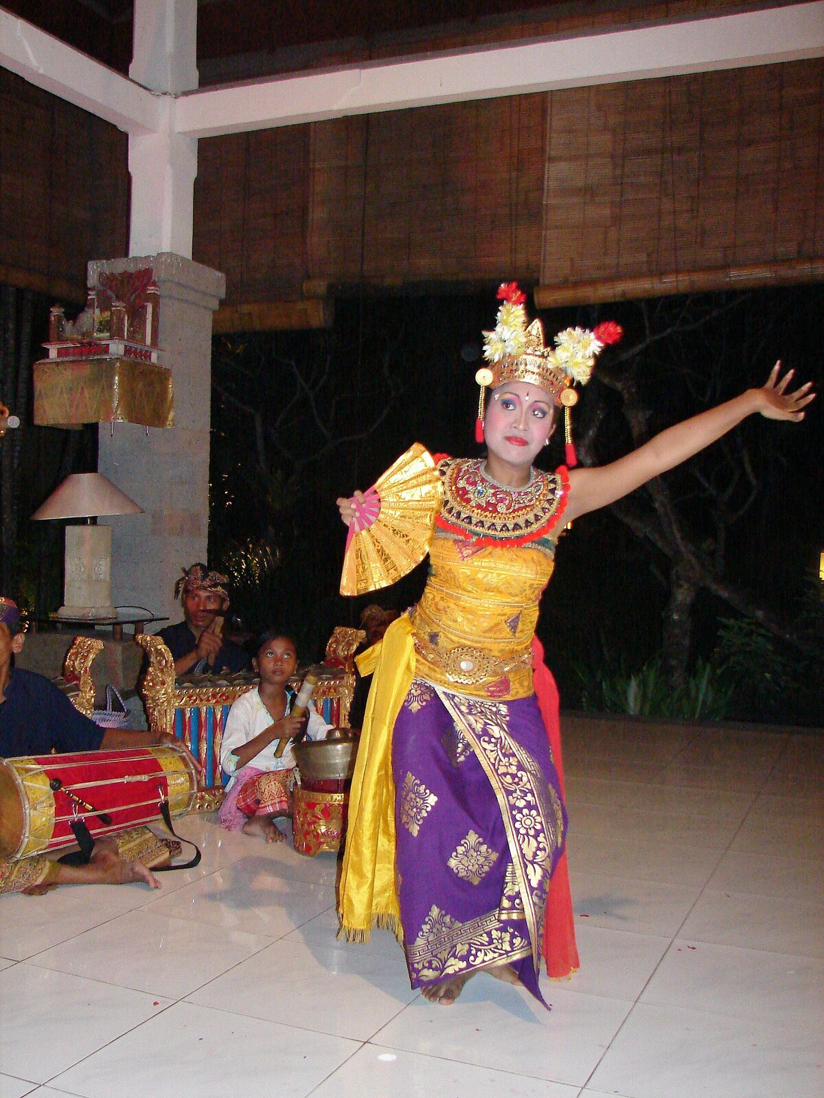 A solo Legong dancer, arms extended, on a courtyard stage.