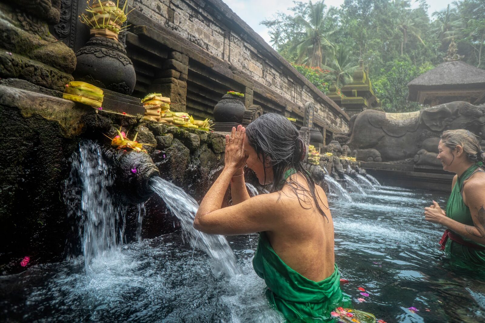 Melukat — bowing head-first under a carved stone spout at a holy spring, letting the water run over you to wash off spiritual impurity.