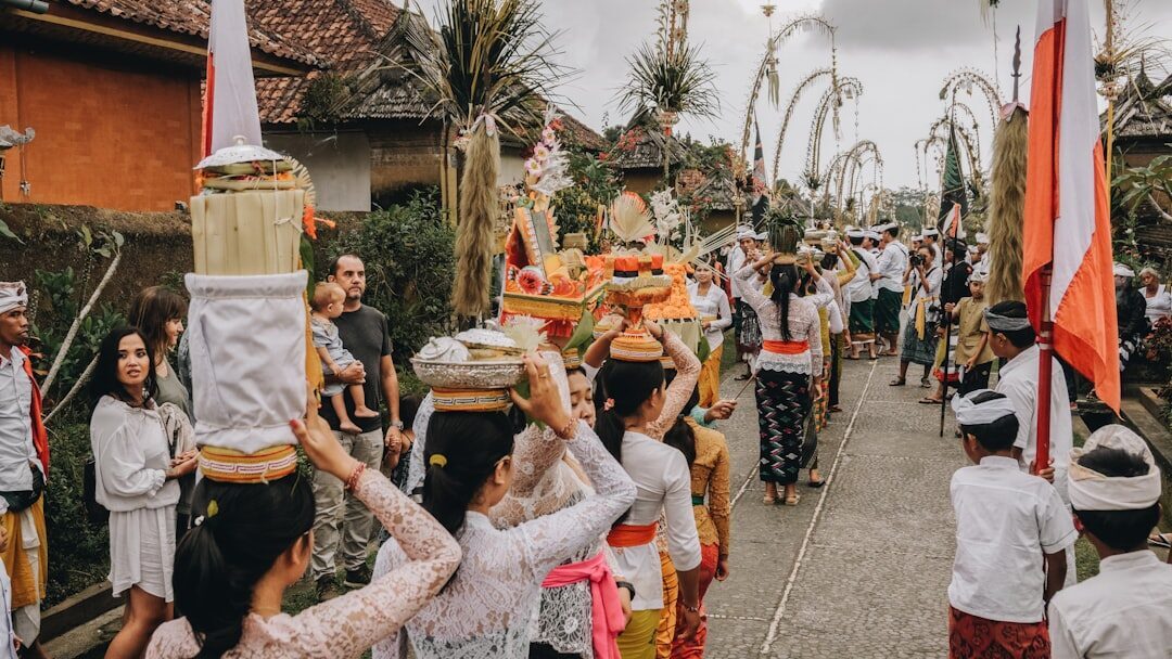 A village procession in white and gold winds toward the temple past the penjor poles.