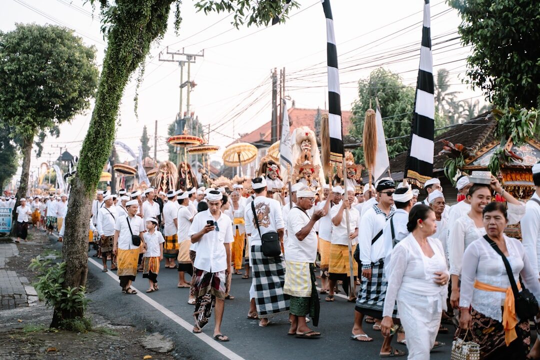 A procession in ceremonial white passing under a street of penjor.