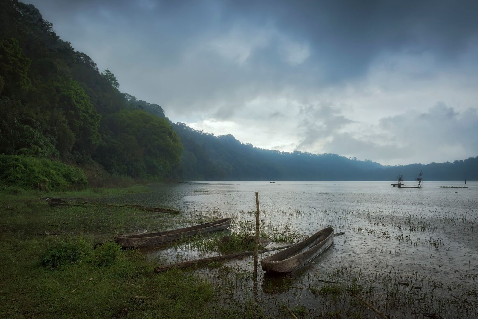 Fishing outriggers drawn up on the shore of Lake Tamblingan, the forested crater rim behind under low cloud.