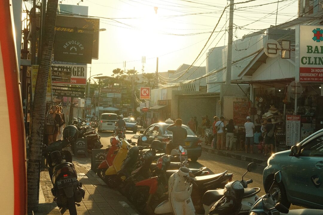 A congested street in warm afternoon light, scooters and cars barely moving past the shops.