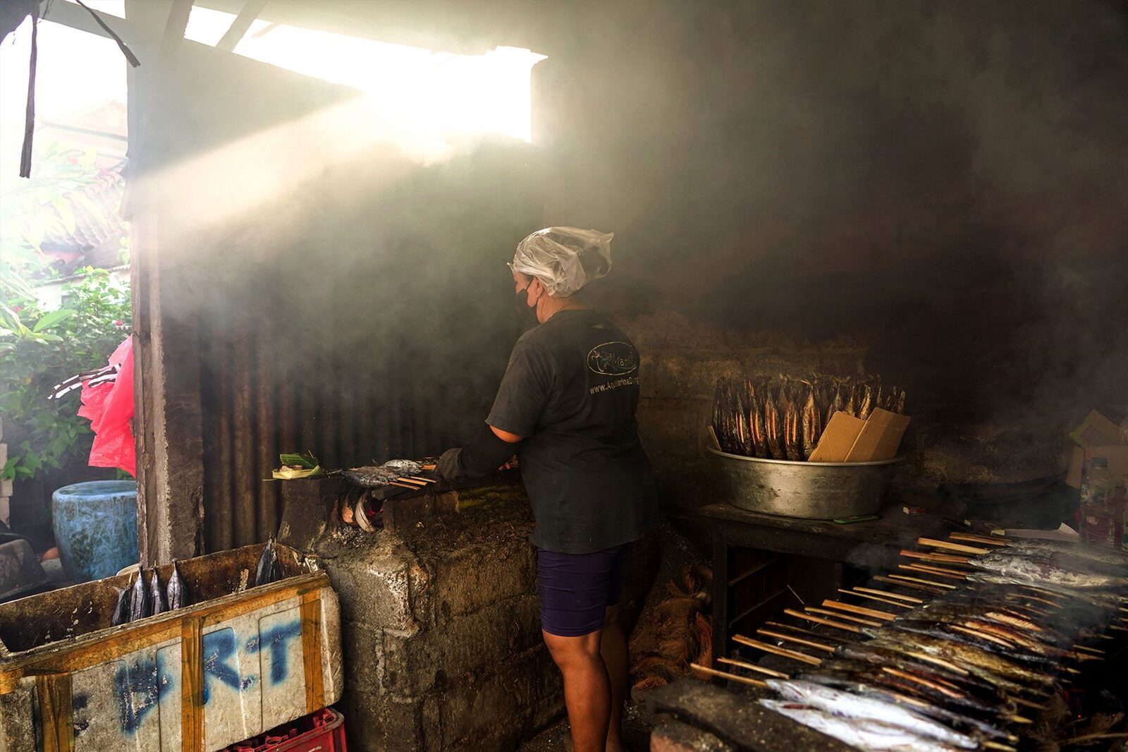 Fish and skewers grilling over coals in a smoky beach kitchen.