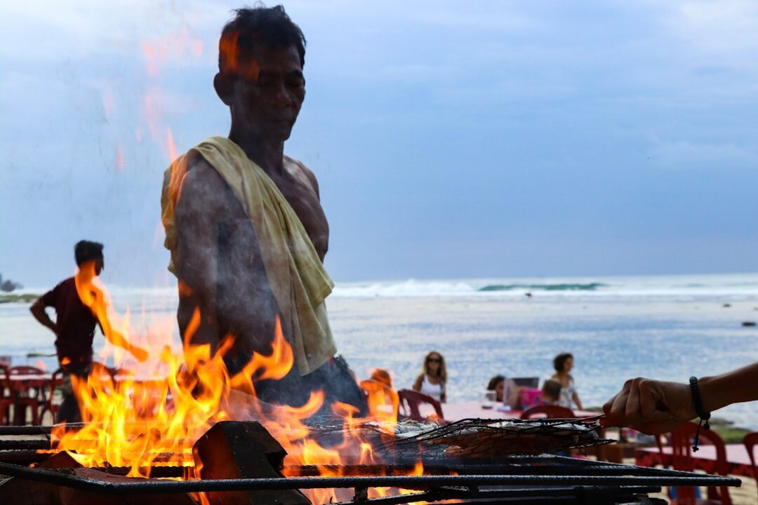 A cook working a roaring open grill on the beach — the smoke and flames of a Jimbaran seafood warung.