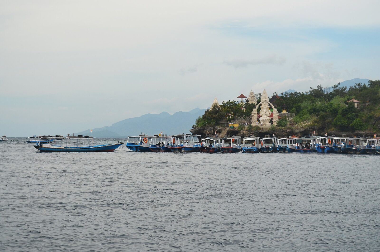 The island's quiet shore, wooden boats moored off a small white temple statue.