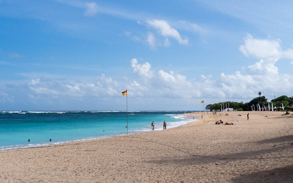 The wide beach with a lifeguard flag flying, water flat behind the offshore reef.
