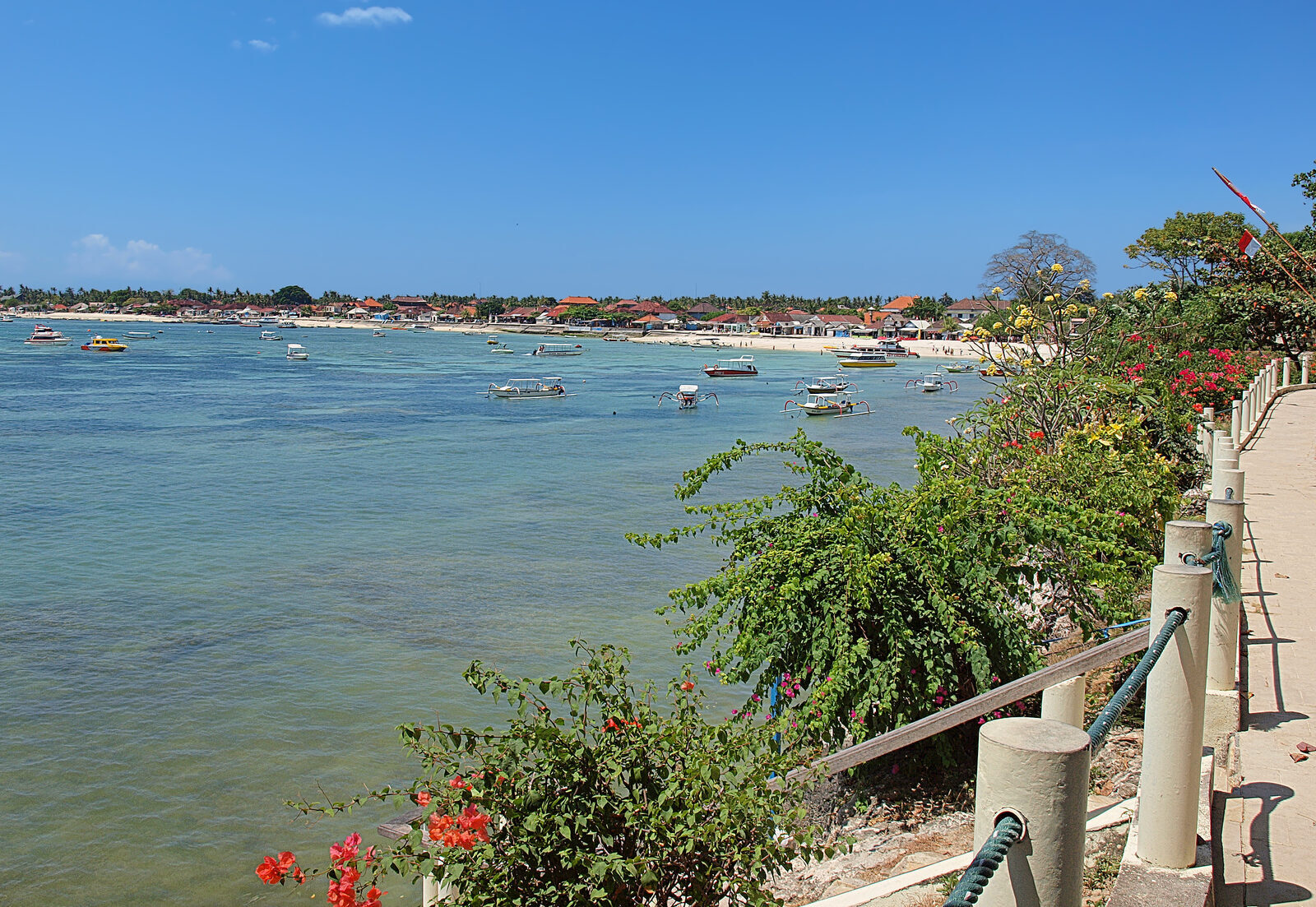 Lembongan's coast from the shore path — reef flats, moored boats and the low green island beyond.