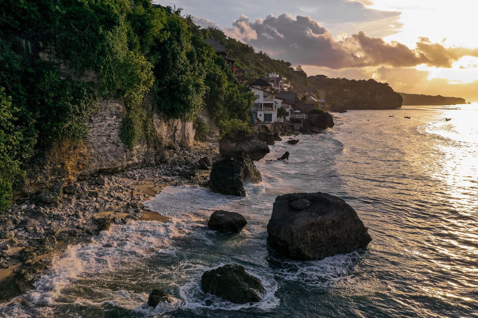 The cliffside cluster of warungs glowing at golden hour, a big rock on the beach.