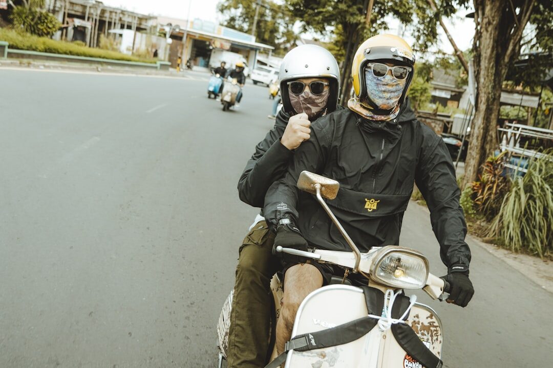 A helmeted couple ride two-up on a scooter down a palm-lined road.