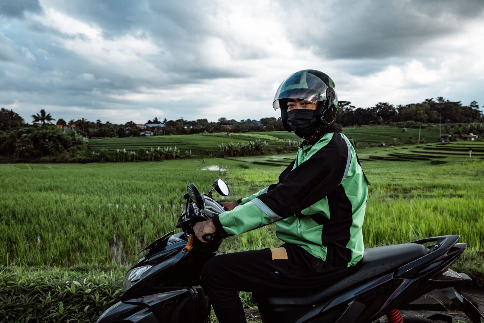 A helmeted driver in a green ride-hailing jacket waits astride his scooter by the rice fields.
