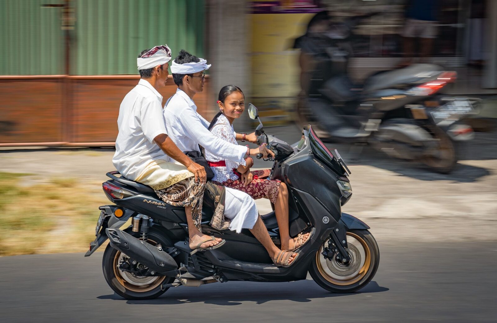 A Balinese family in ceremonial white rides through traffic, motion streaking past.