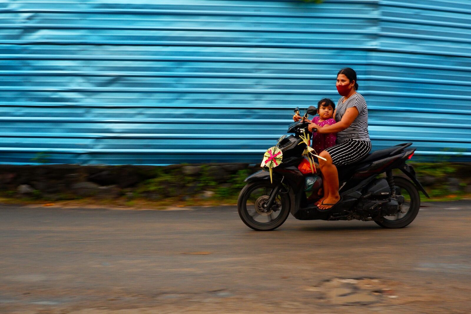 A mother and child ride a scooter past a painted wall — everyday Bali on two wheels.