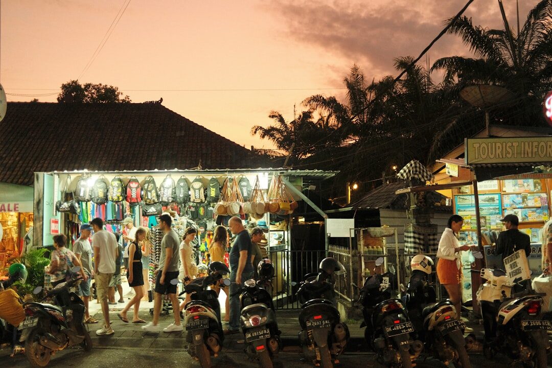 A market street at dusk, scooters parked and passing under the warung lights.