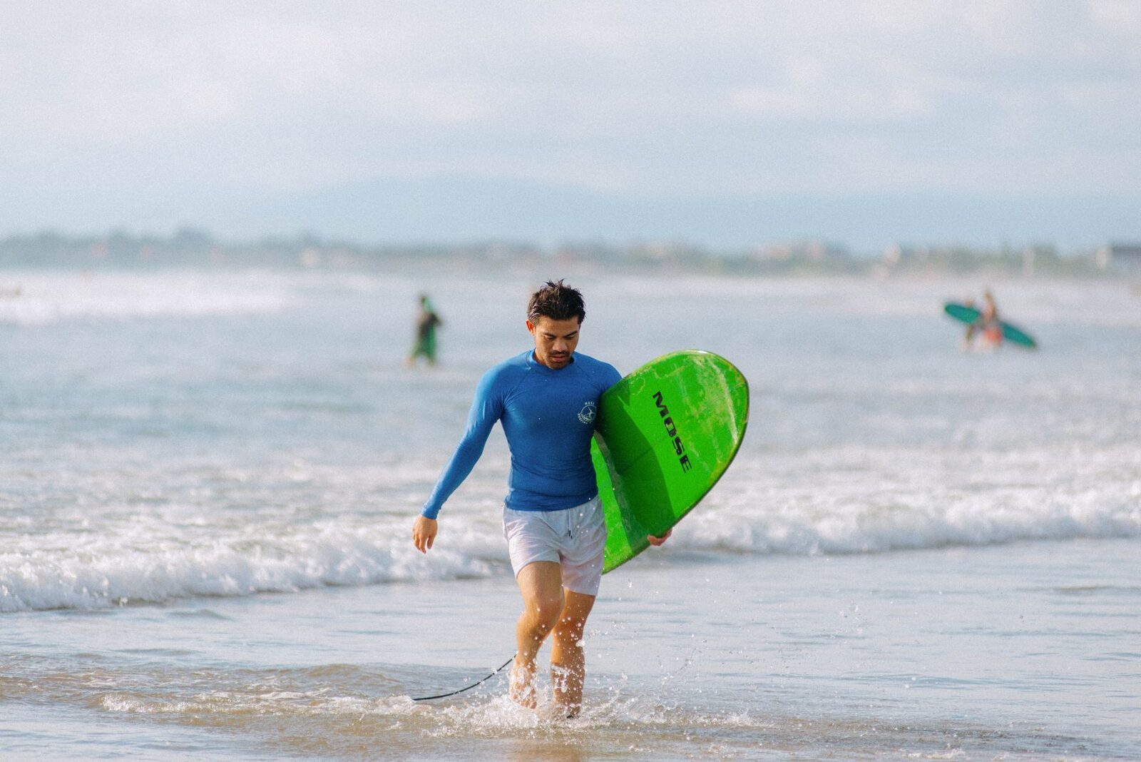 A surfer walking out with a soft-top board at a mellow beach break — sand under you, not reef.