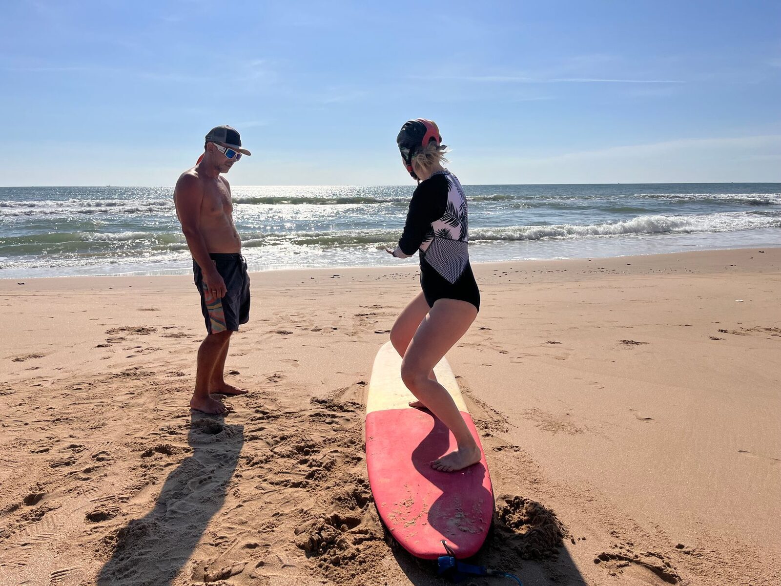 An instructor and student with a soft-top board on the sand — the real value of a lesson is someone reading the tide, rip and peak for you.