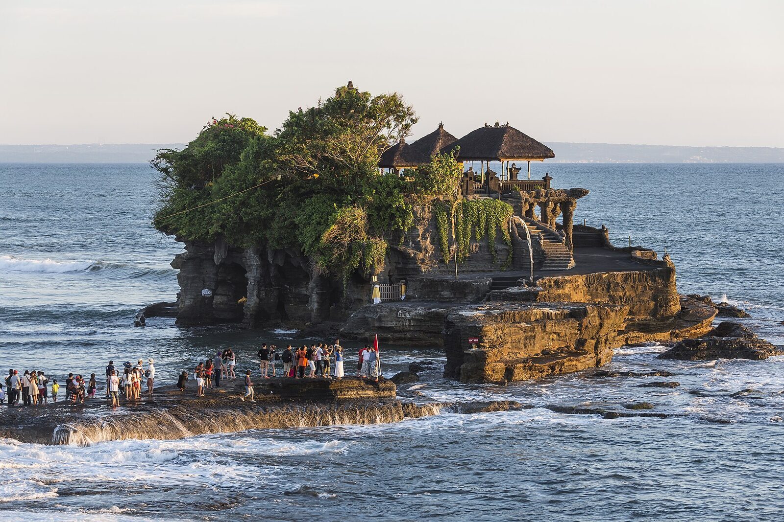 Pura Tanah Lot on its rock, a crowd gathered at the base and surf breaking around it — the classic view from the shore.