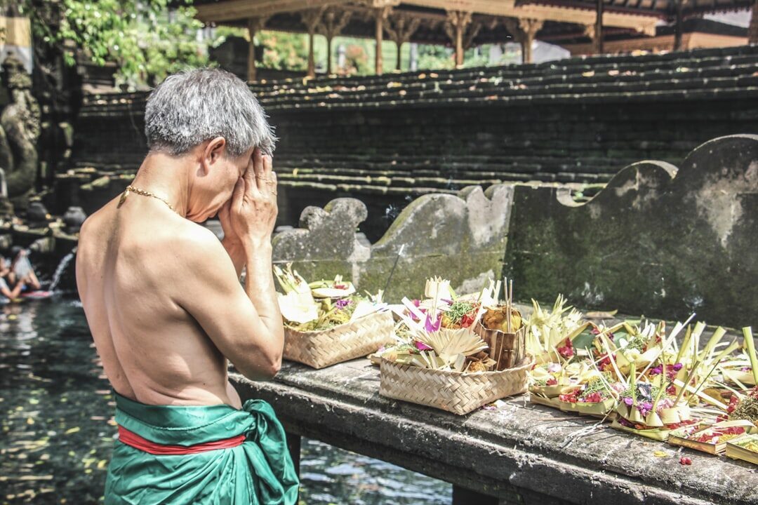 Melukat up close — head bowed and hands raised at a spout, an offering set on the ledge above.