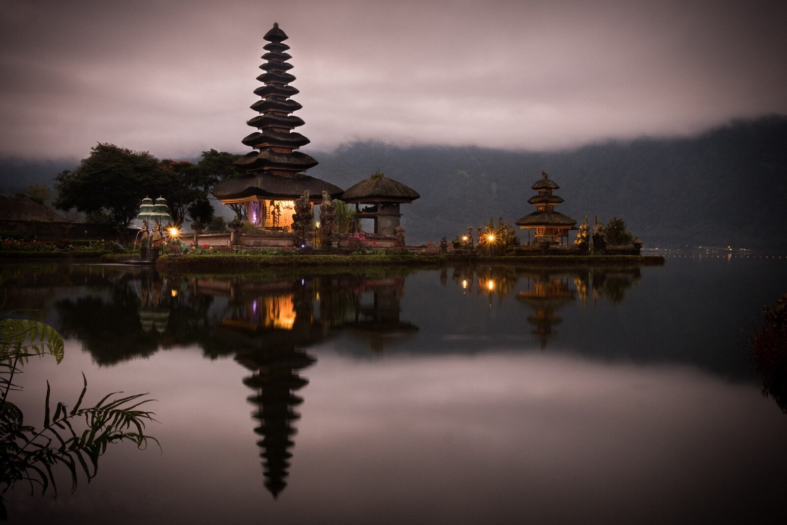 First light at Ulun Danu Beratan: warm lamplight in the shrine and a near-perfect mirror in the still dawn water — the reason to arrive at opening.