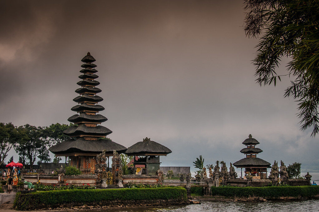 The wider complex — meru, stupa and gateway — under a moody Bedugul sky.